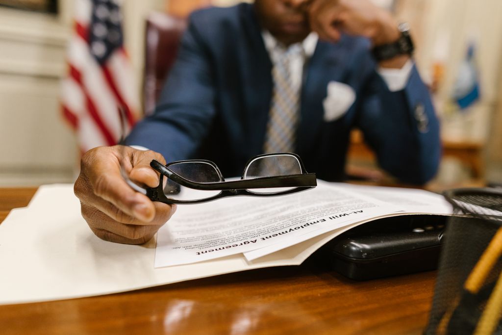 Business professional contemplating a contract, wearing a suit, seated at a desk with documents and eyeglasses.